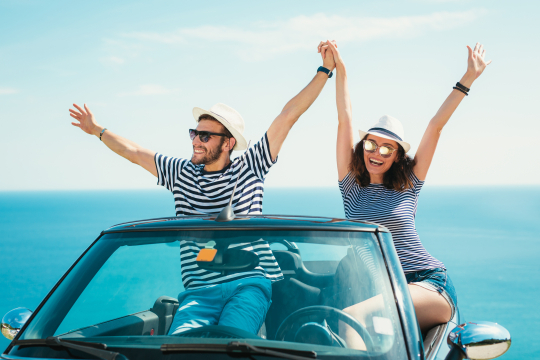 Pärchen sitzt auf Rückbank eines Cabriolets und streckt die Arme in den Himmel, im Hintergrund das Meer